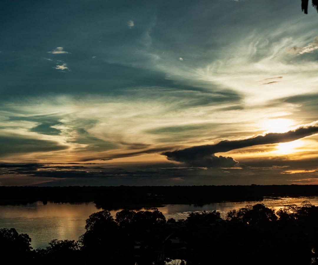 Atardecer sobre un río con reflejos dorados, rodeado de árboles oscuros y un cielo con nubes dramáticas iluminadas.