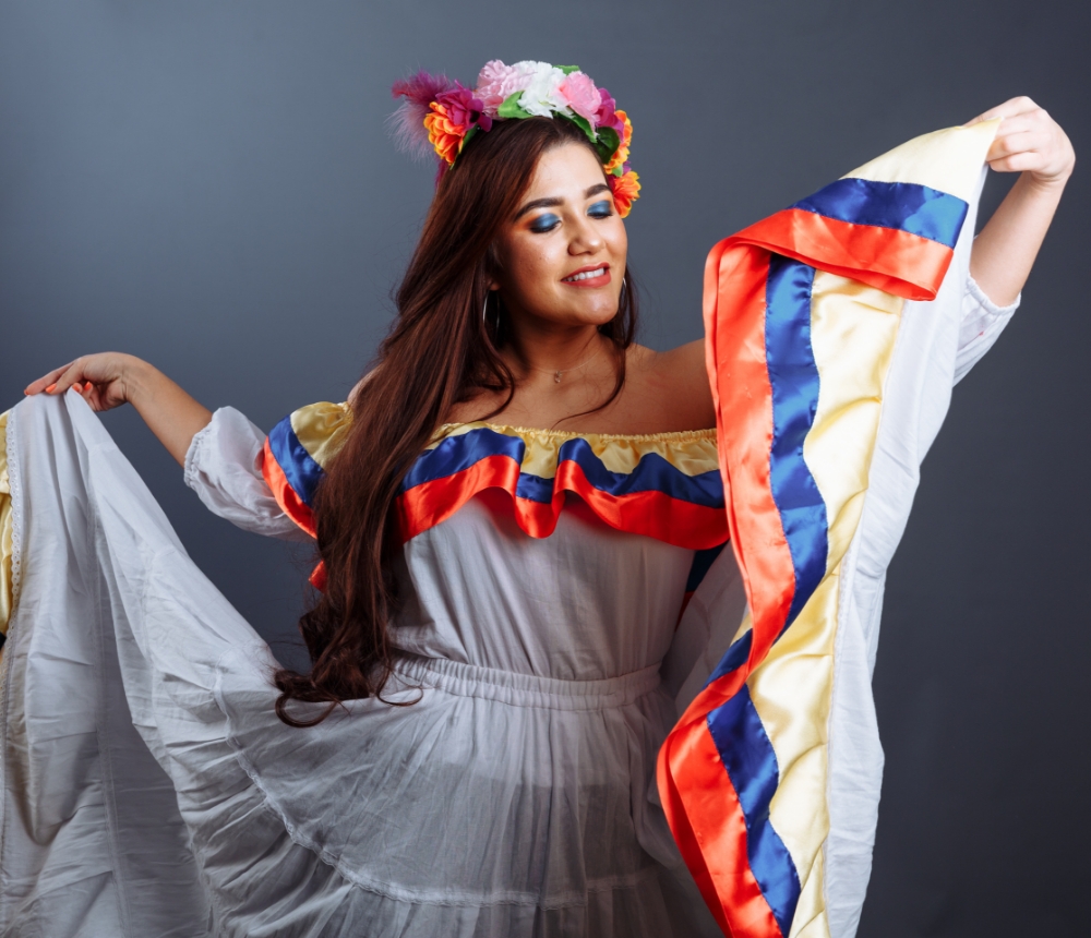 Woman dressed in traditional white folk costume with details in yellow, blue and red, holding the skirt and with a wreath on her head.