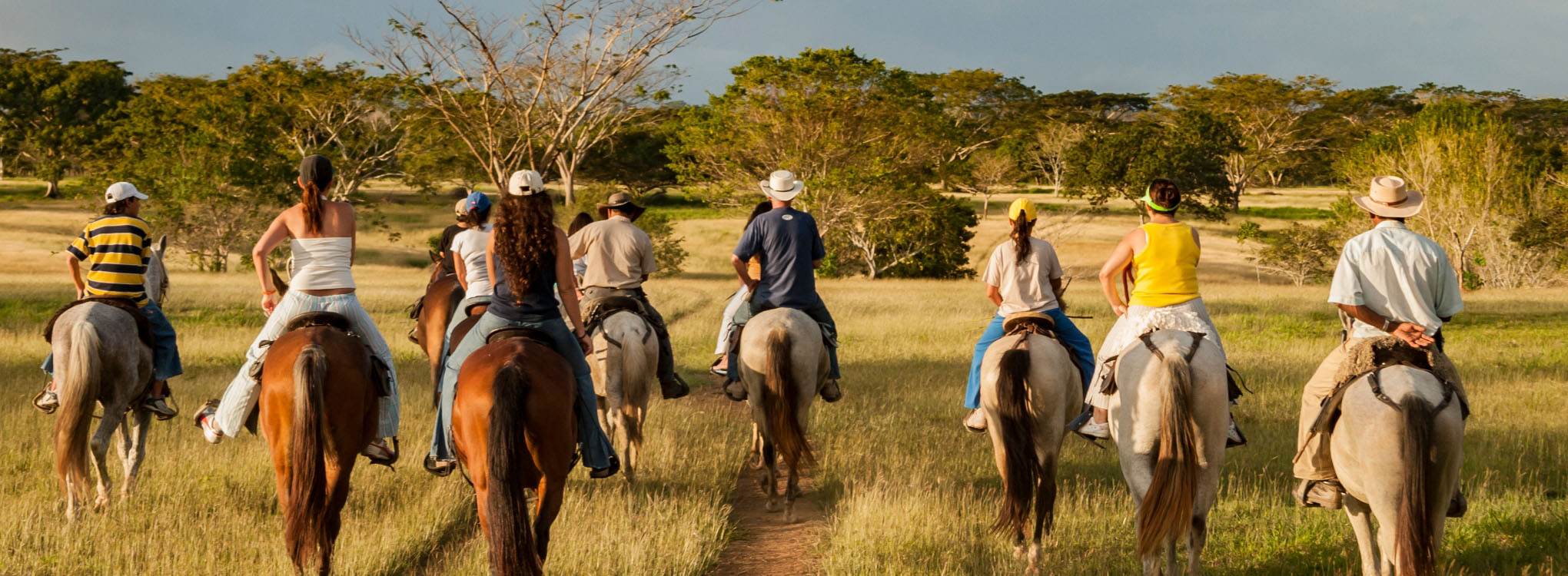 Grupo de personas montando a caballo en un paisaje rural con pastizales y árboles bajo un cielo parcialmente nublado.