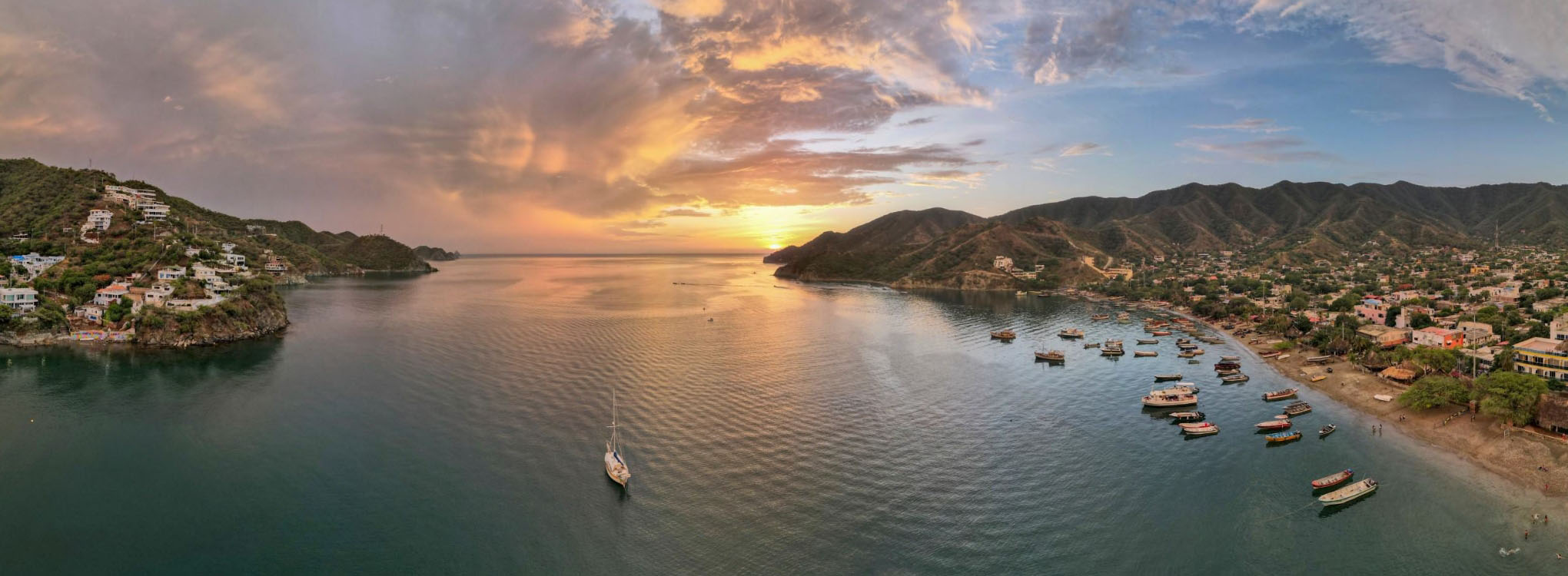 Vista panorámica al atardecer de una bahía con colinas, casas en la ladera, barcos en el agua y un cielo coloreado con tonos naranjas y rosas.