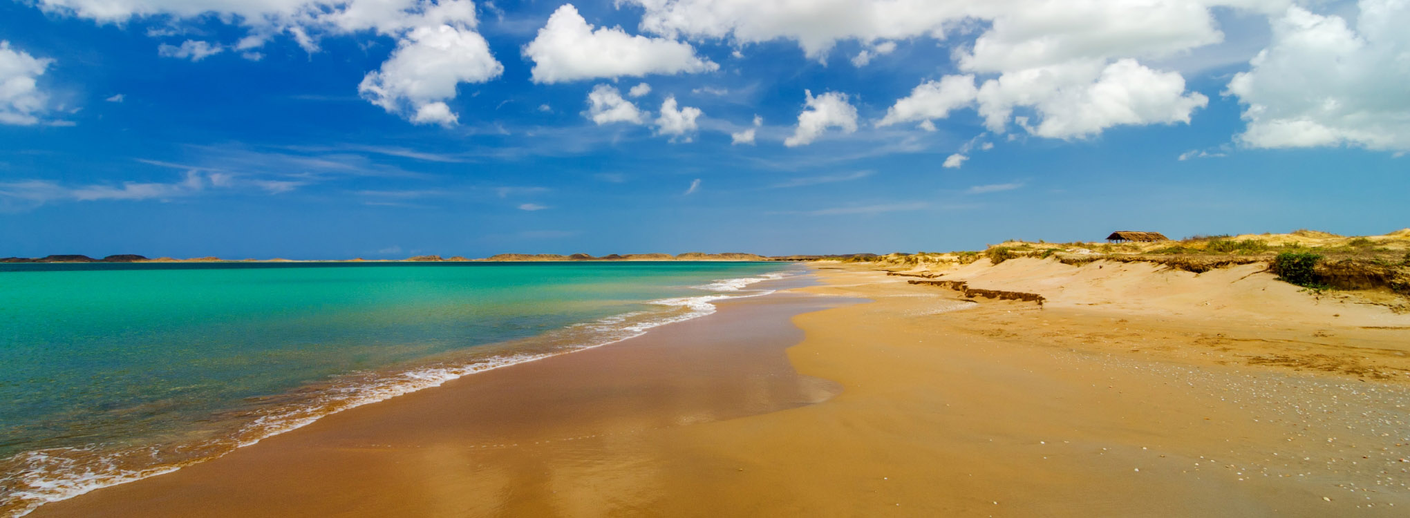 Playa de aguas cristalinas y arena dorada, con un cielo azul brillante decorado con nubes blancas, en un entorno natural y sereno.