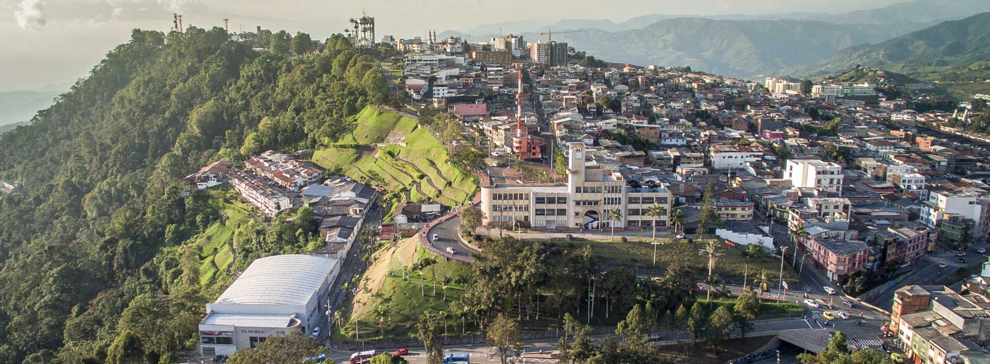 Vista panorámica de Manizales, mostrando calles, rotondas, áreas verdes y edificios con montañas al fondo.