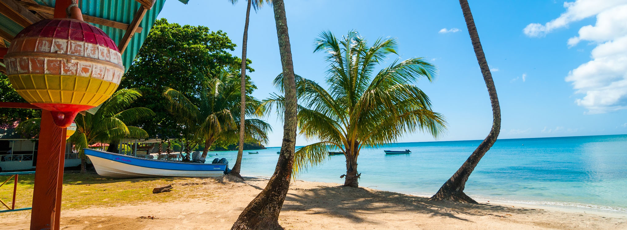 Playa tropical con palmeras inclinadas, una lancha en la orilla y aguas cristalinas, bajo un cielo azul despejado con nubes blancas.