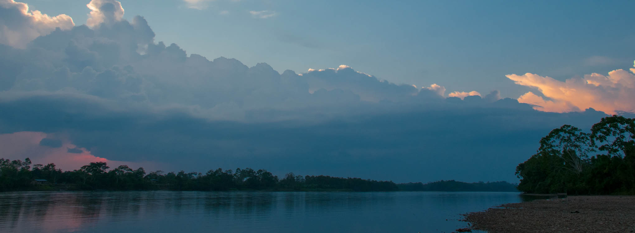 Paisaje al atardecer con un río tranquilo, árboles en la orilla y un cielo con nubes iluminadas.