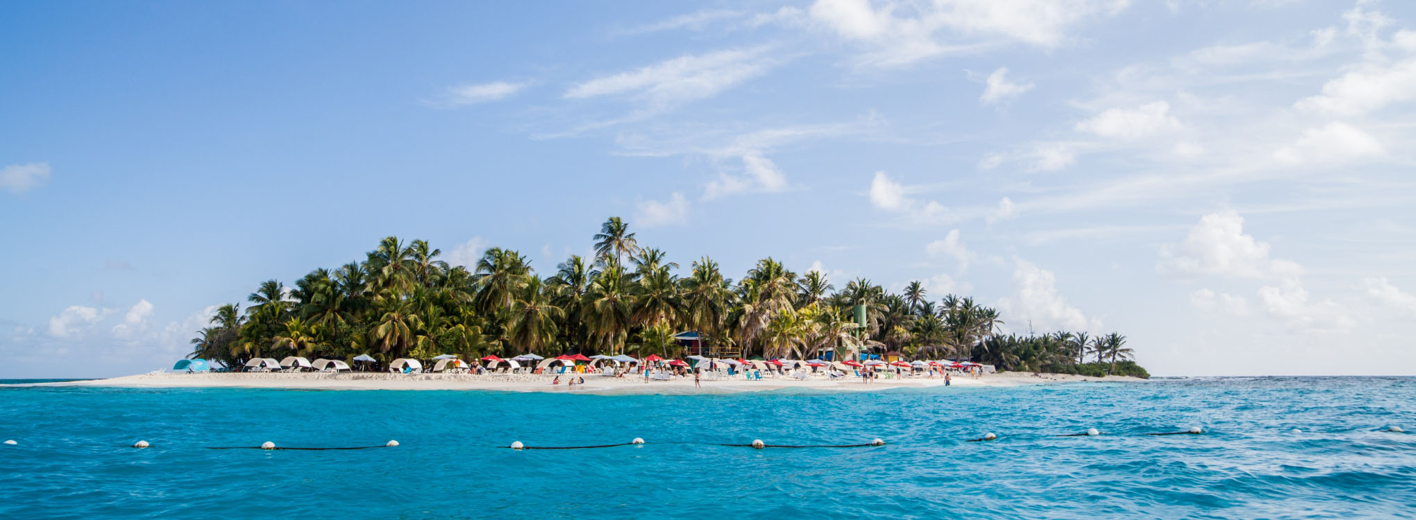 Isla tropical rodeada por aguas cristalinas de color turquesa, con una playa de arena blanca, palmeras y sombrillas para los visitantes