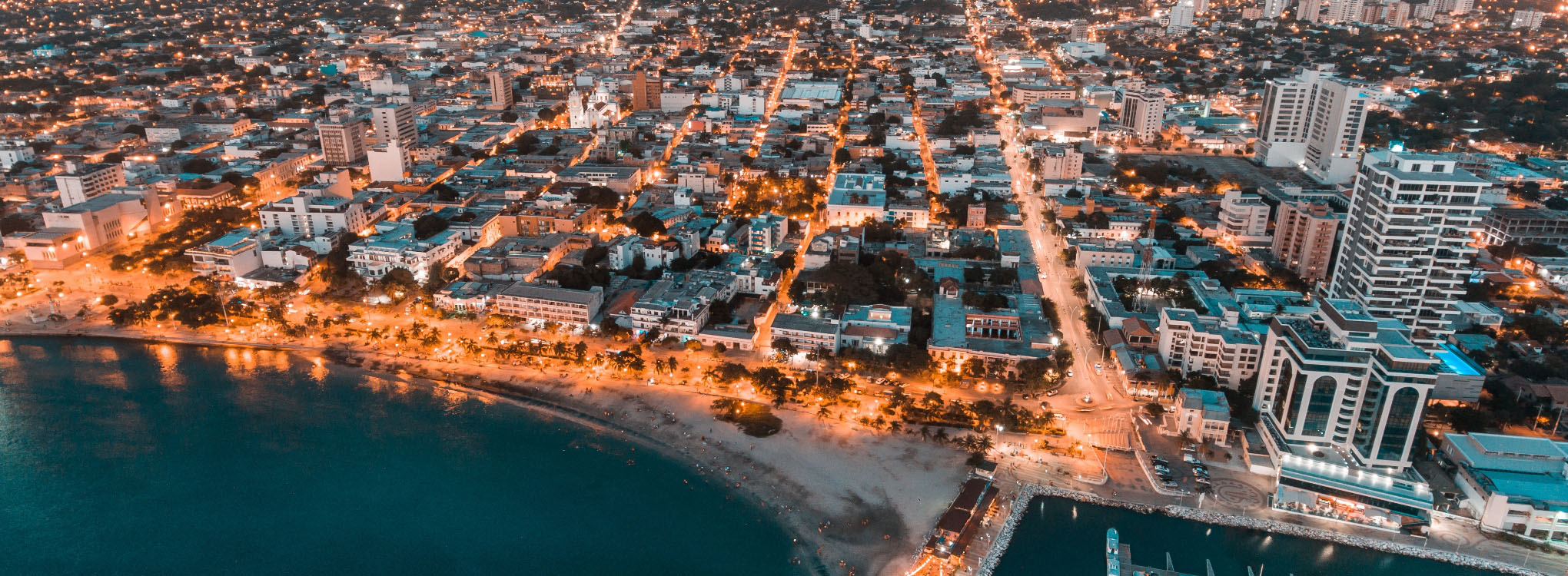 Vista aérea nocturna de una ciudad costera iluminada, con edificios, calles bien trazadas y una playa bordeada por luces junto al mar.