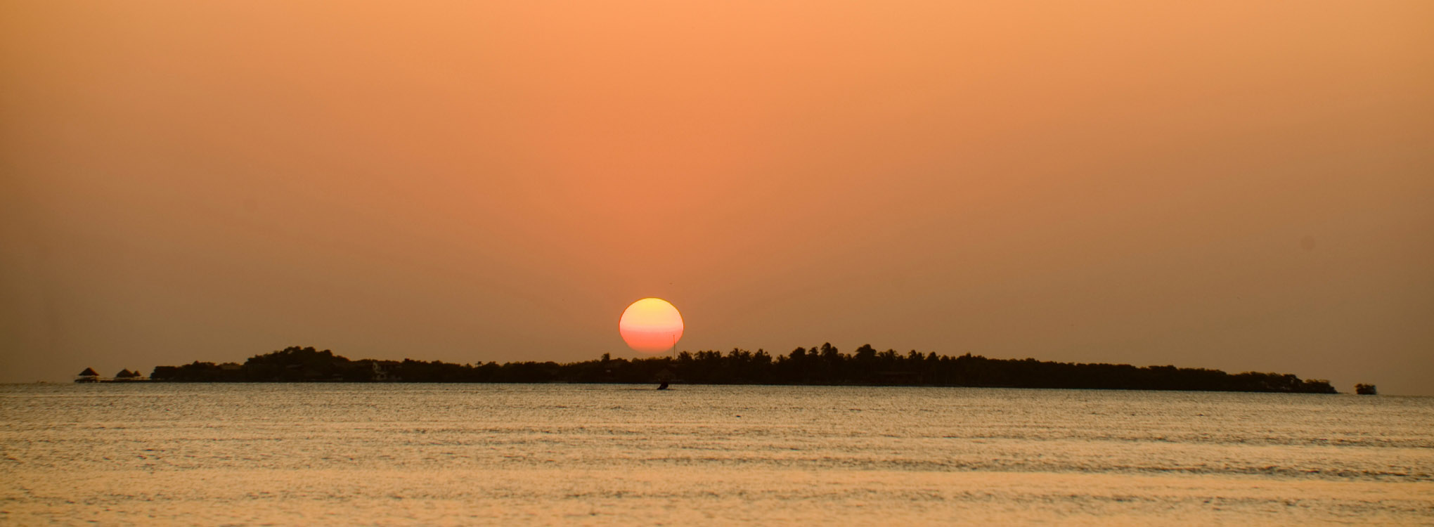 Atardecer con el sol rojo en el horizonte, reflejado en el agua tranquila, con una isla cubierta de árboles en la distancia.