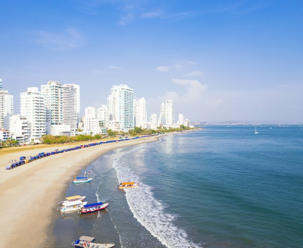 Vista panorámica de una playa con arena dorada, barcos pequeños en la orilla y edificios altos frente al mar, bajo un cielo despejado.