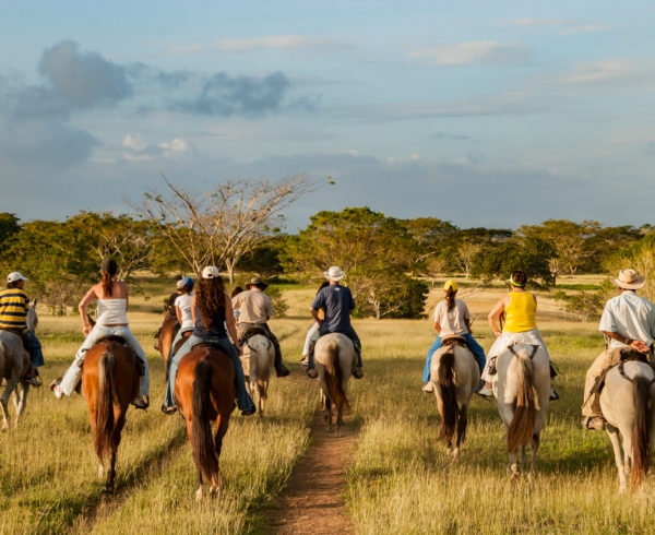 Grupo de personas montando a caballo en un paisaje rural con pastizales y árboles bajo un cielo parcialmente nublado.