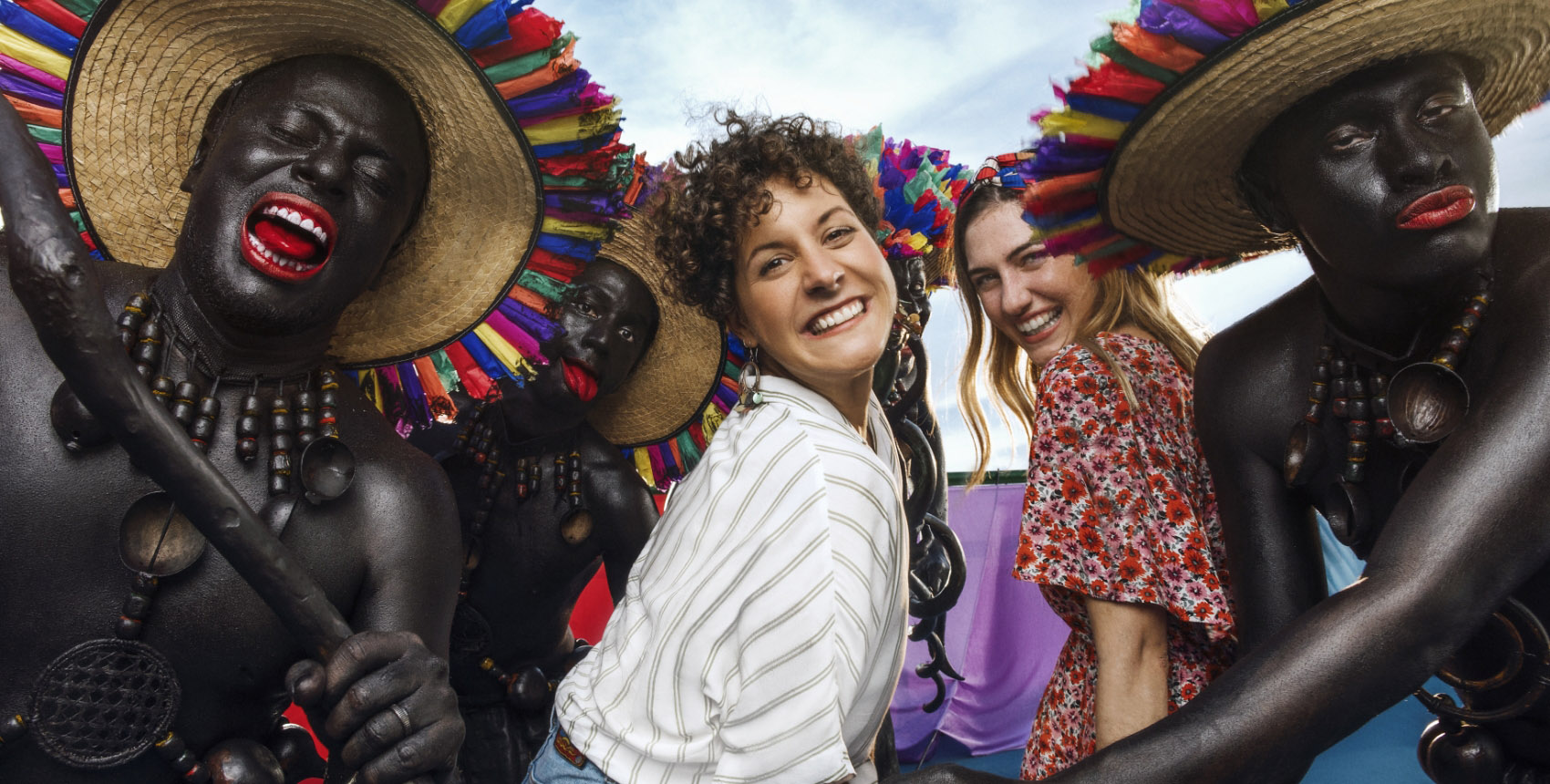 Grupo de personas en una celebración cultural, con personajes pintados de negro y sombreros de colores, acompañados por dos mujeres sonrientes.