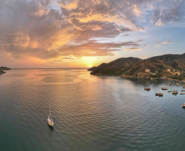 Vista panorámica al atardecer de una bahía con colinas, casas en la ladera, barcos en el agua y un cielo coloreado con tonos naranjas y rosas.