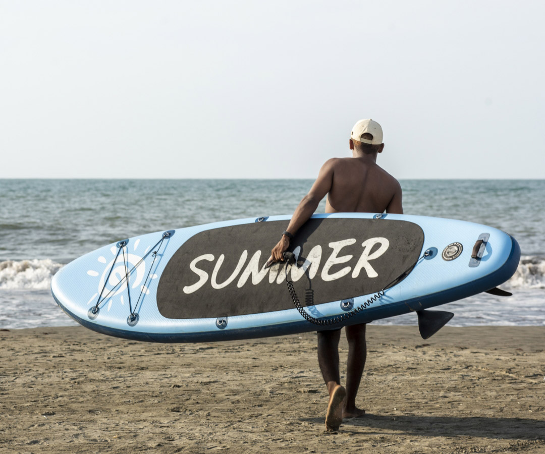 Hombre caminando hacia el mar en la playa con una tabla de paddle surf azul con la palabra «Verano» escrita en ella.