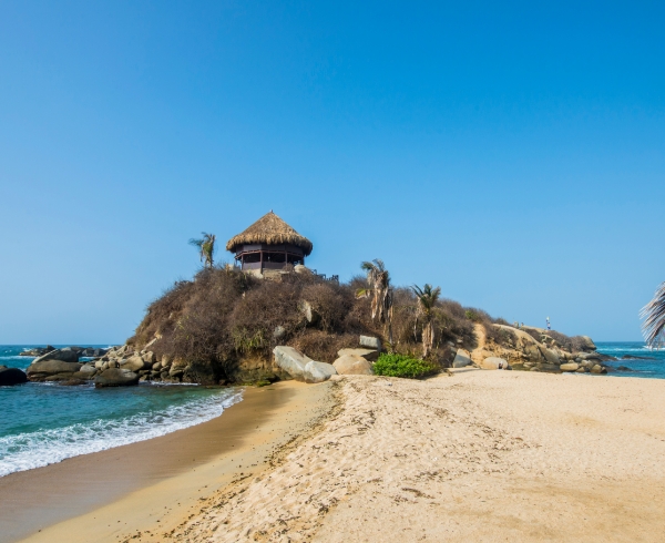 Vista de una playa con arena dorada y un cabo rocoso que alberga una cabaña con techo de palma, rodeada de vegetación, bajo un cielo despejado.