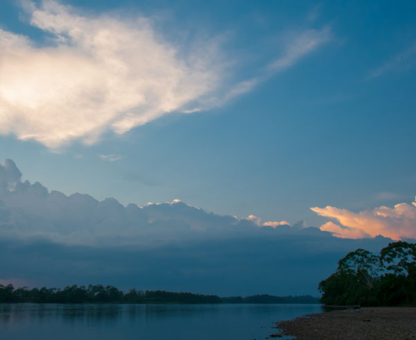 Paisaje al atardecer con un río tranquilo, árboles en la orilla y un cielo con nubes iluminadas.