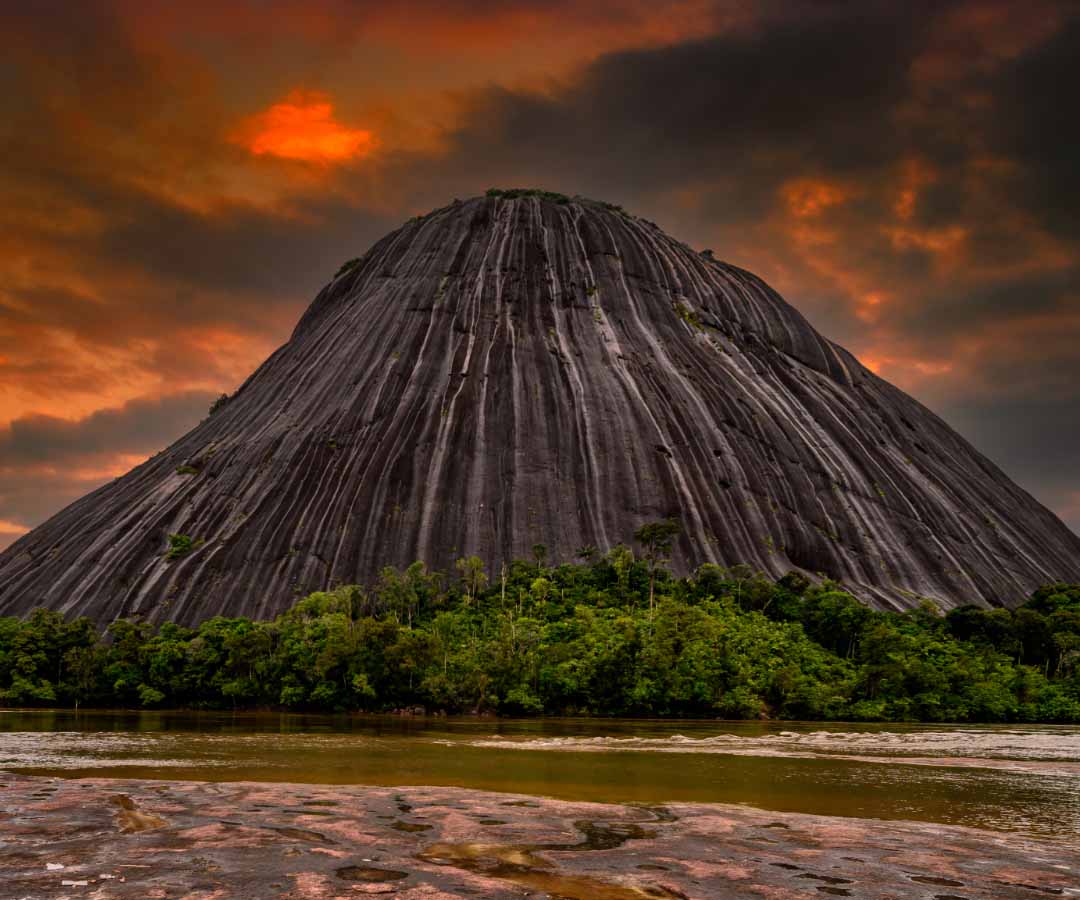 Imponente montaña rocosa rodeada de vegetación y un río, con un cielo dramático teñido de tonos anaranjados.