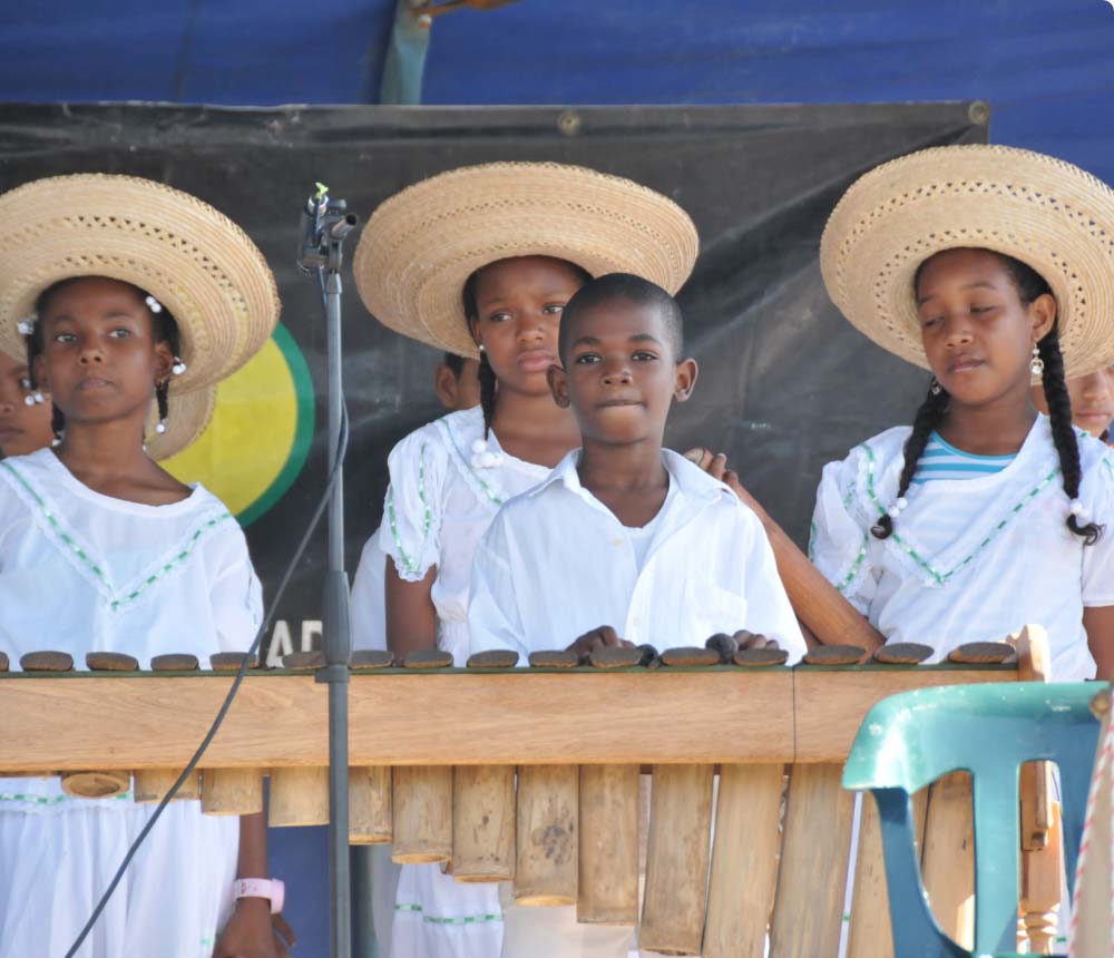 Grupo de niños vestidos con trajes tradicionales blancos y sombreros de paja, tocando un instrumento musical.