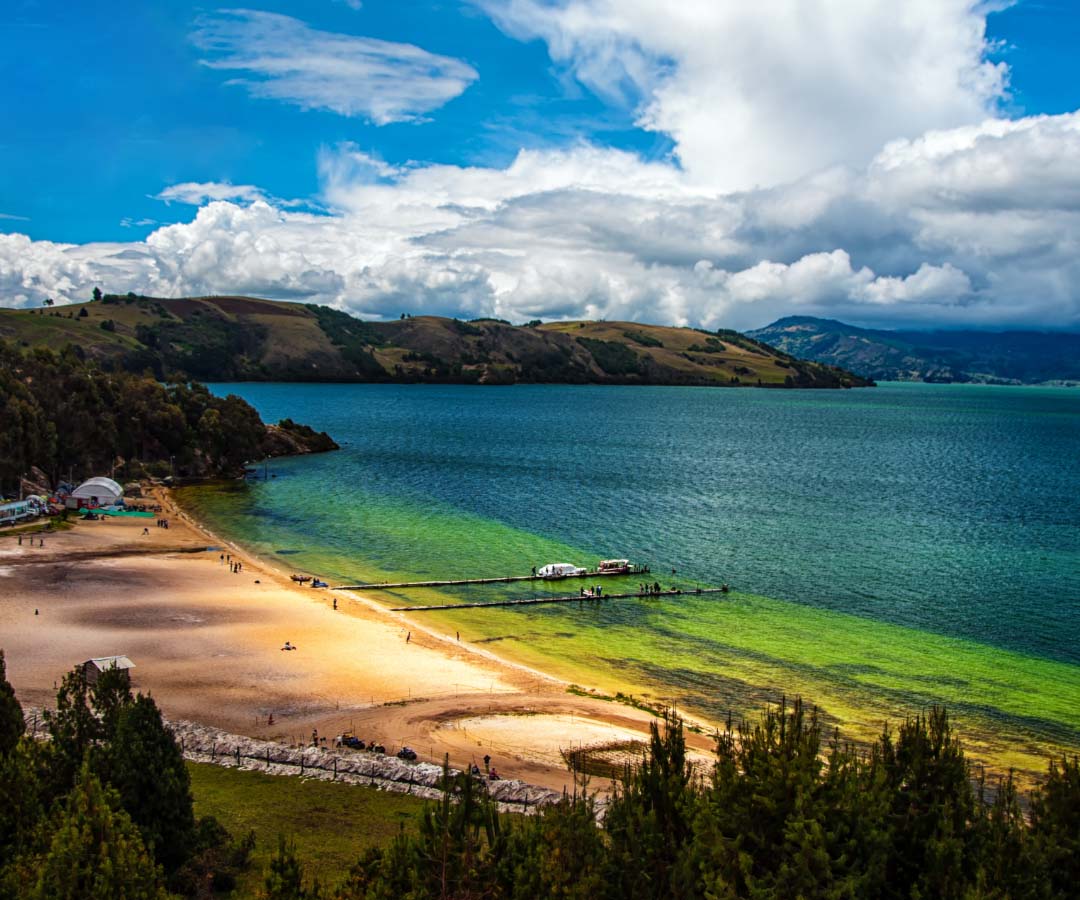 Vista panorámica de una playa junto a un lago de aguas cristalinas, rodeado de colinas verdes.