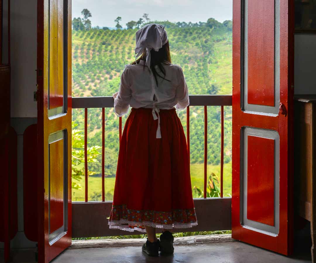 Mujer con traje típico del Eje Cafetero, de espaldas, observando un paisaje de cafetales.