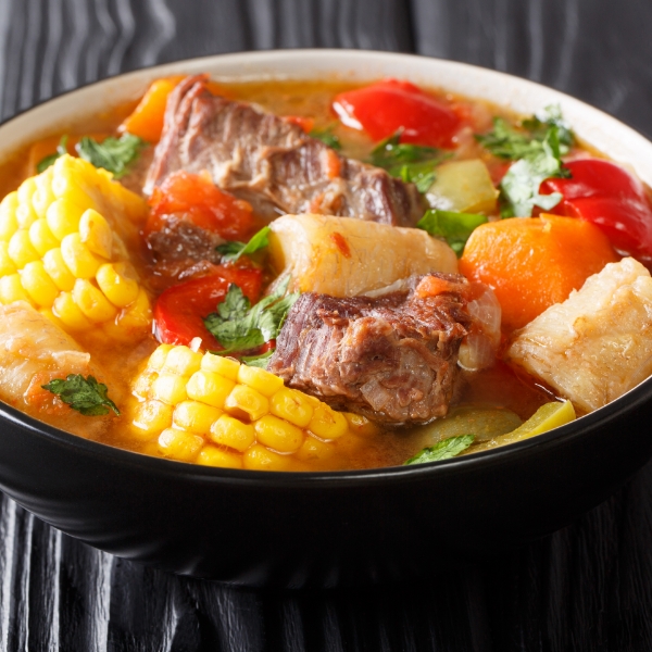 Sancocho, a traditional soup with meat, corn, yucca, plantain, carrot and cilantro, served in a dark bowl.