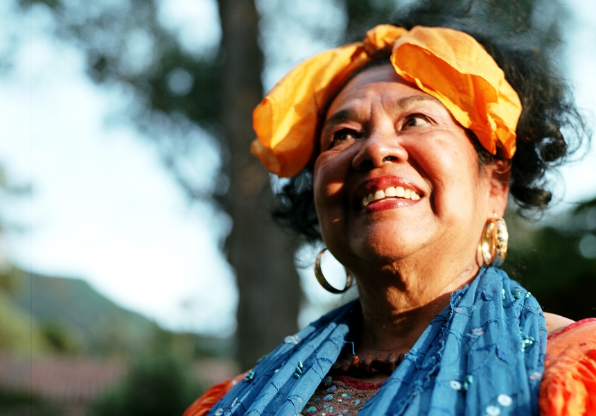 Totó la Momposina sonriendo con un pañuelo amarillo en la cabeza, grandes pendientes y un pañuelo azul, al aire libre. 