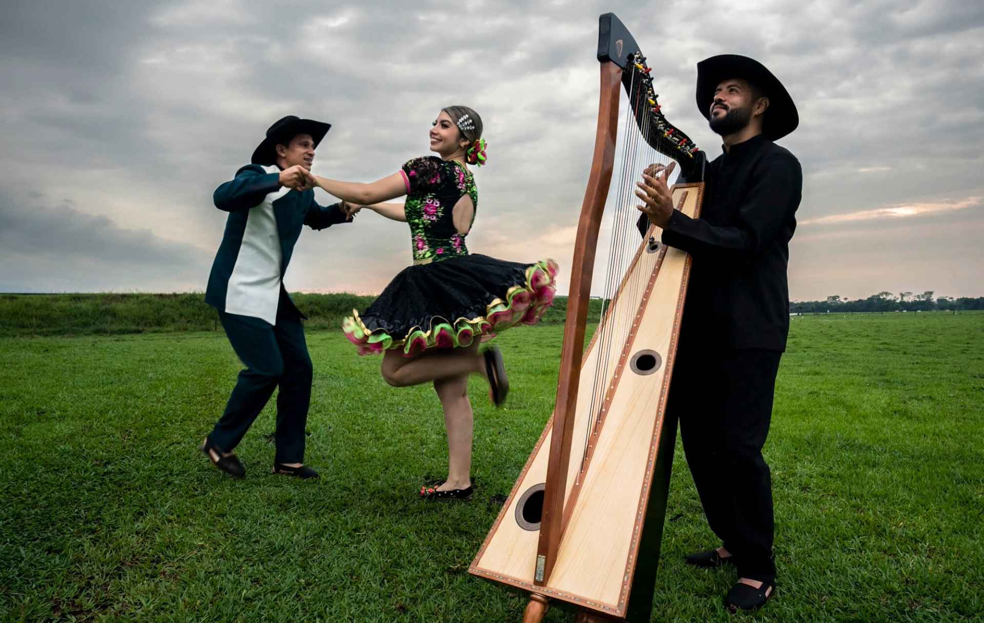 Pareja bailando joropo mientras un músico toca el arpa, en un paisaje llanero al aire libre.