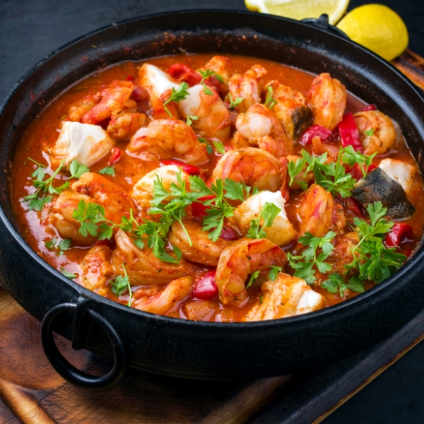 Cazuela de marisco con gambas, pescado, pimientos y hierbas frescas, servida en un cuenco de barro oscuro.