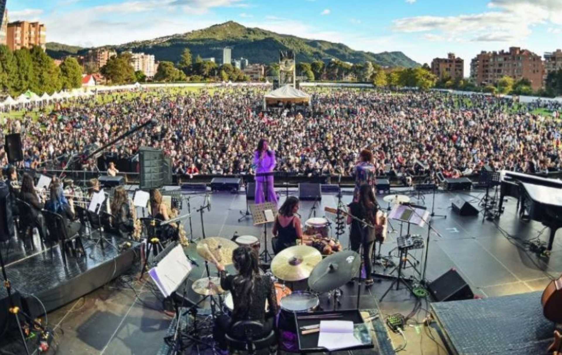 Escenario de un concierto al aire libre, con una orquesta en primer plano y una multitud numerosa disfrutando del evento