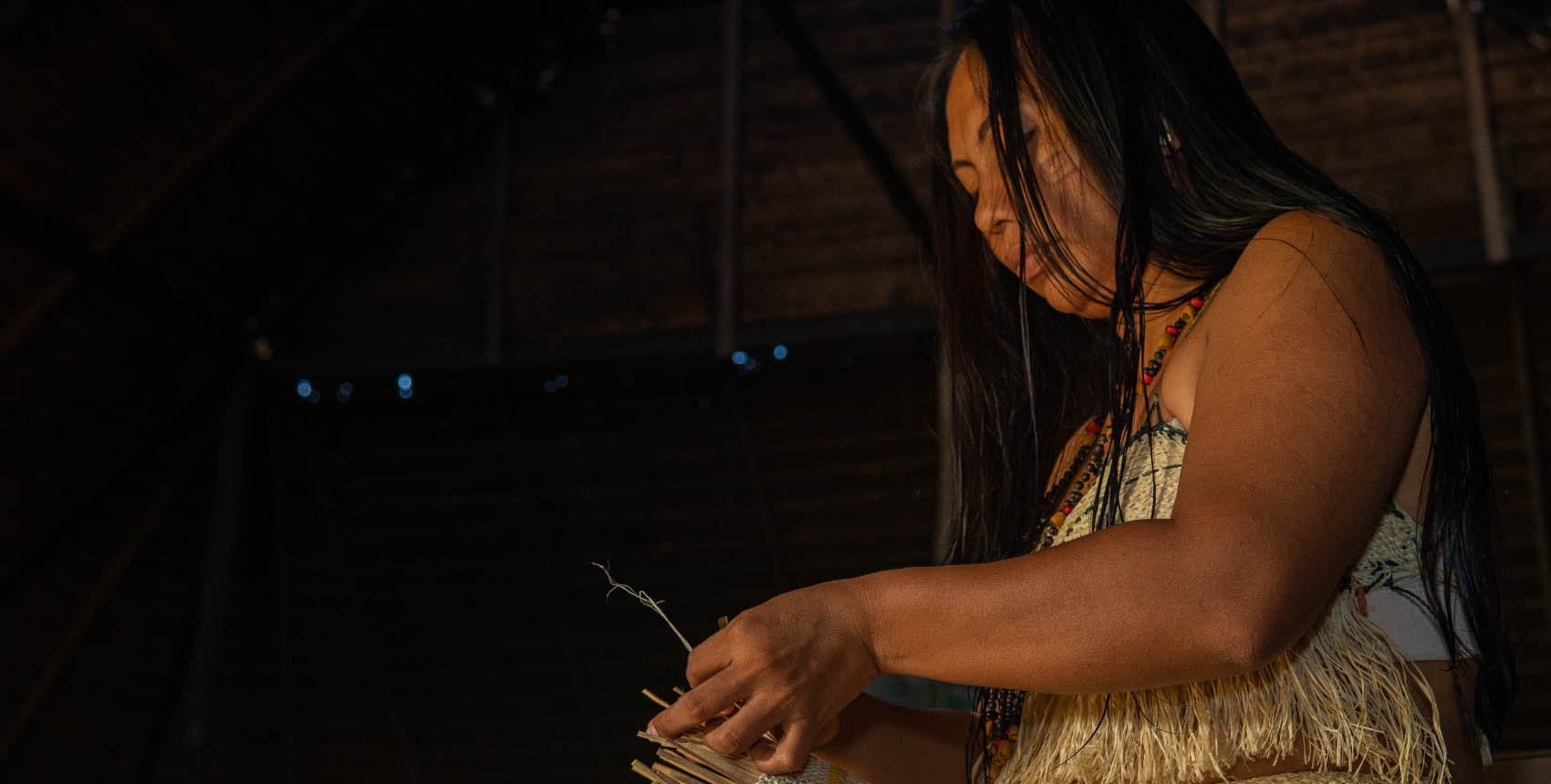 Mujer indígena en el interior de una choza, sosteniendo materiales naturales en sus manos.