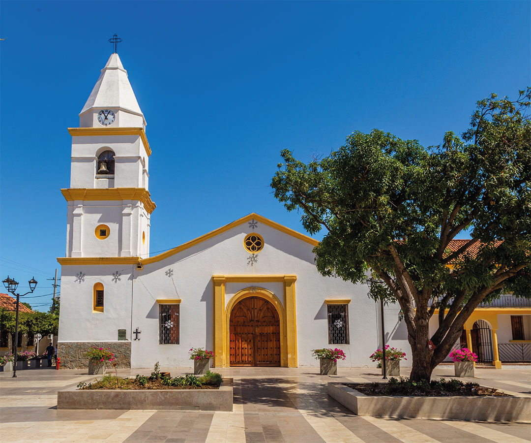 Iglesia colonial en Valledupar, con fachada blanca y detalles amarillos, rodeada de árboles y flores.