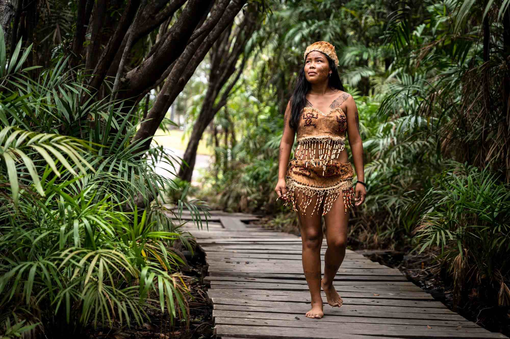 Mujer indígena caminando descalza por un sendero de madera rodeado de vegetación.