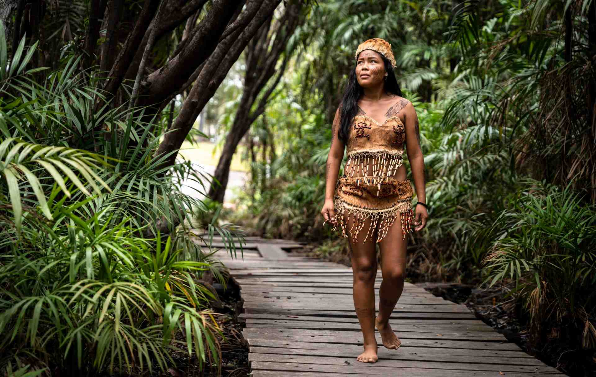 Mujer indígena caminando descalza sobre un sendero de madera rodeado de una exuberante vegetación.