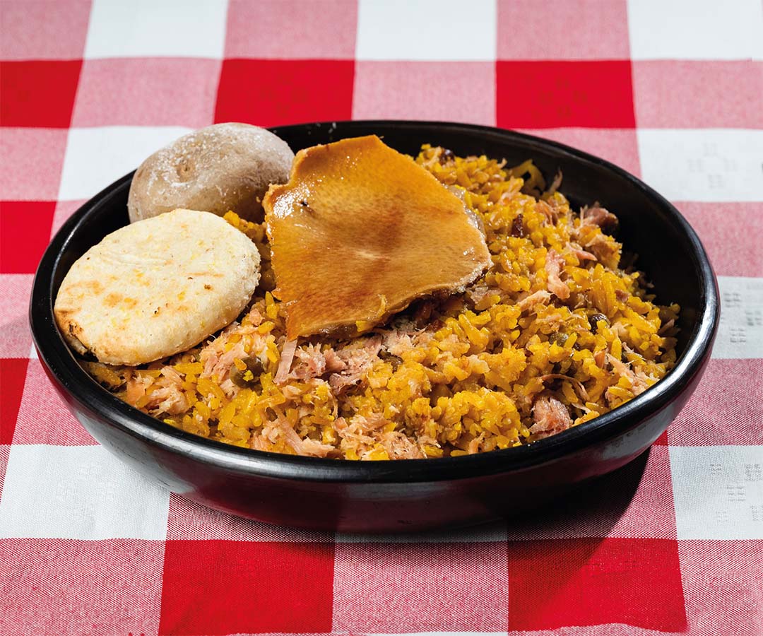 Plato típico de Lechona tolimense, servido con arroz, carne de cerdo, arepa y papa, sobre un mantel de cuadros rojos y blancos.