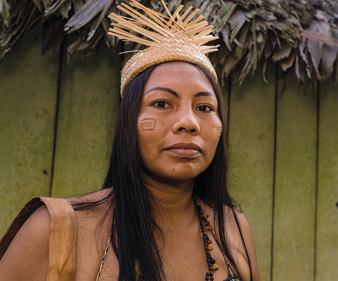 Mujer indígena con vestimenta tradicional y corona artesanal de paja, de pie frente a una pared de madera.