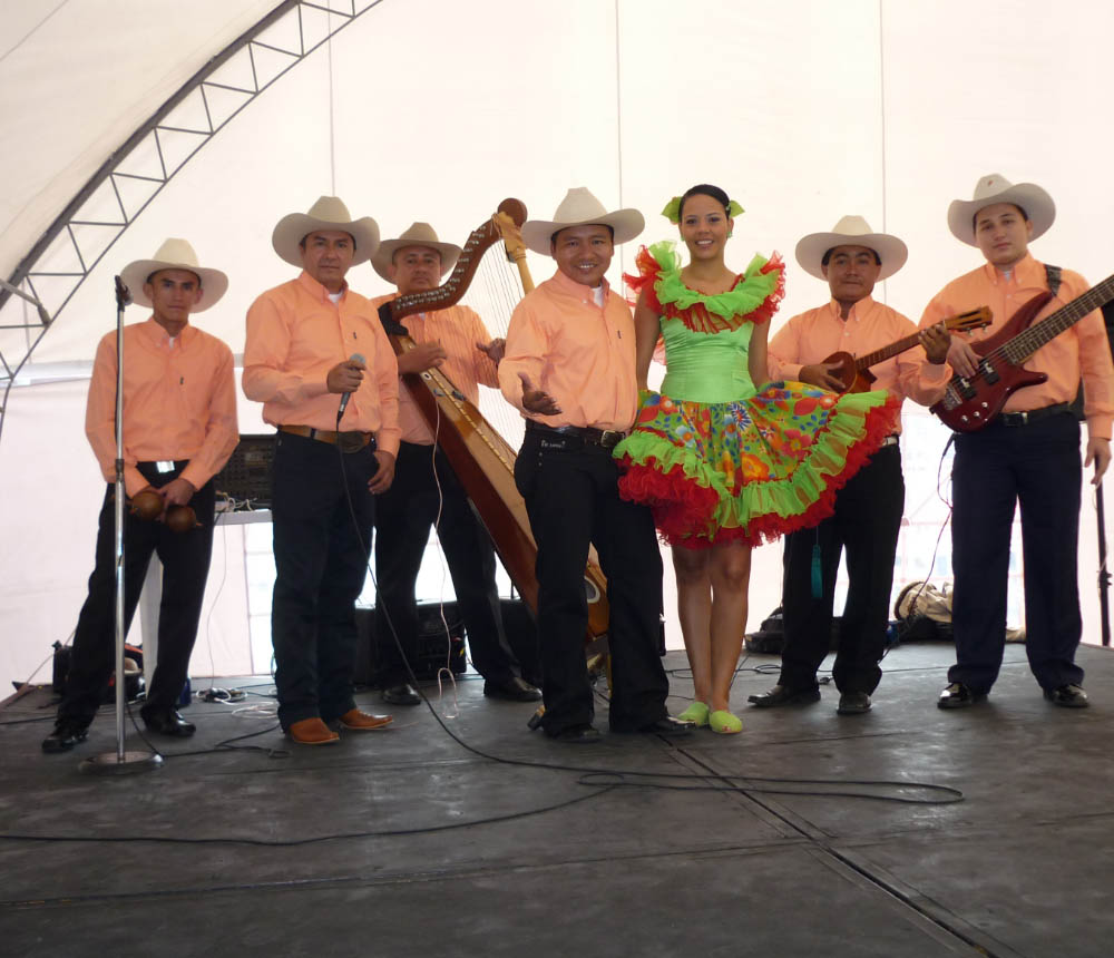 Grupo musical tradicional posando en un escenario, con músicos vestidos con camisas naranjas y sombreros.