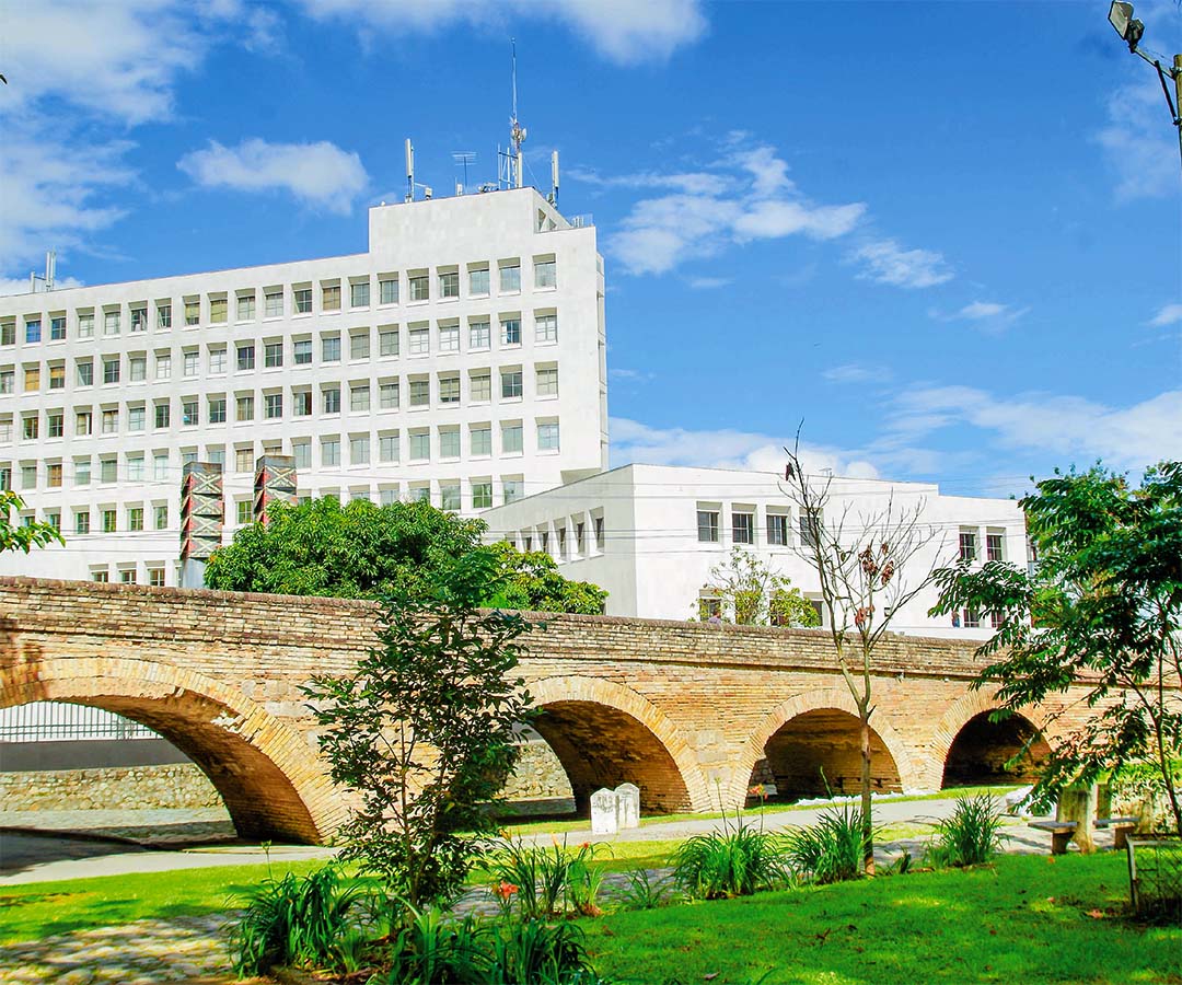 Puente colonial de Popayán rodeado de vegetación, con un edificio moderno al fondo y un cielo despejado.