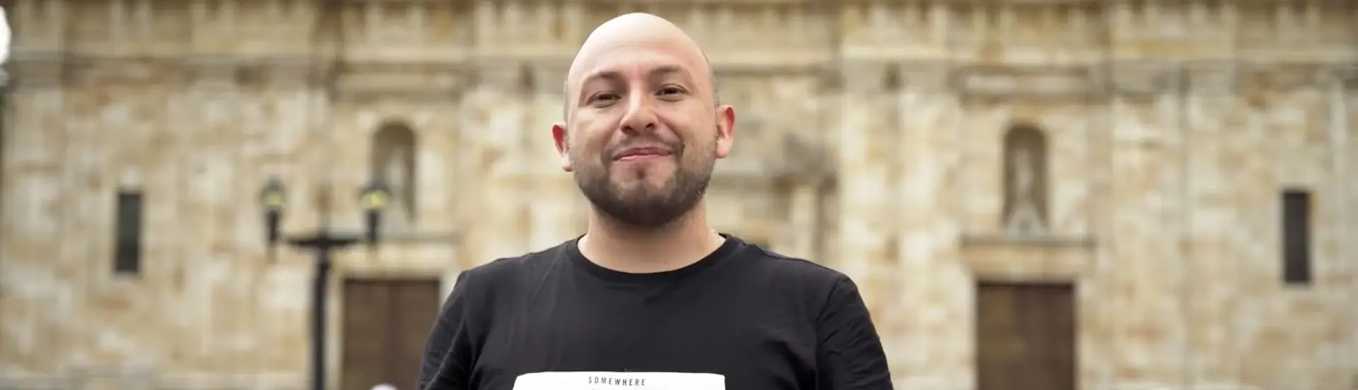 Hombre calvo con camiseta negra sonriendo frente a edificio histórico en Colombia
