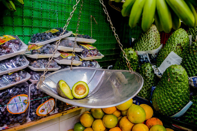 plaza de mercado, pesa con un aguacate hass partido sobre ella con guanabana y naranjas de fondo, recetas de jugos naturales