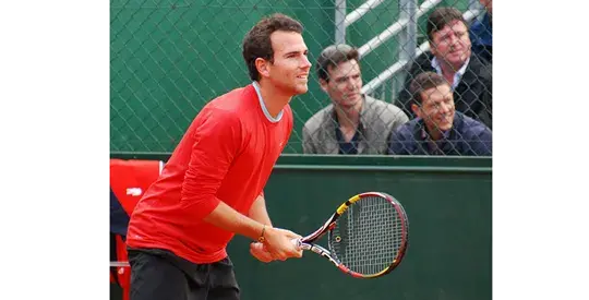 Tenista Adrian Mannarino en la cancha de arcilla, listo para recibir la pelota, con público de fondo.