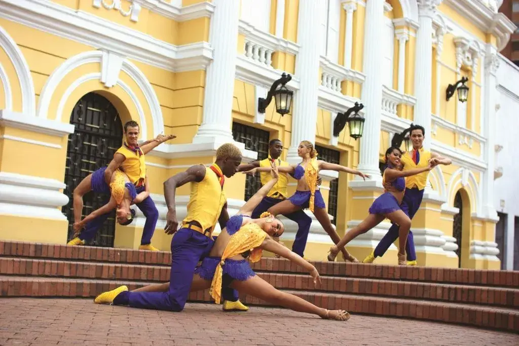 Bailarines de salsa con trajes amarillos y morados frente a edificio colonial.