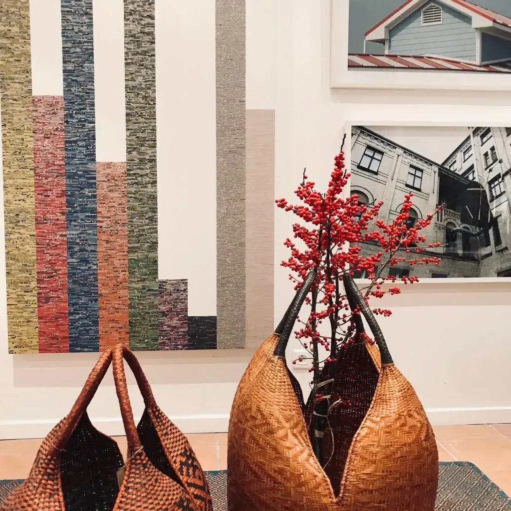 Bolsos de fibra tejida y rama roja ante tapiz abstracto y fotos.