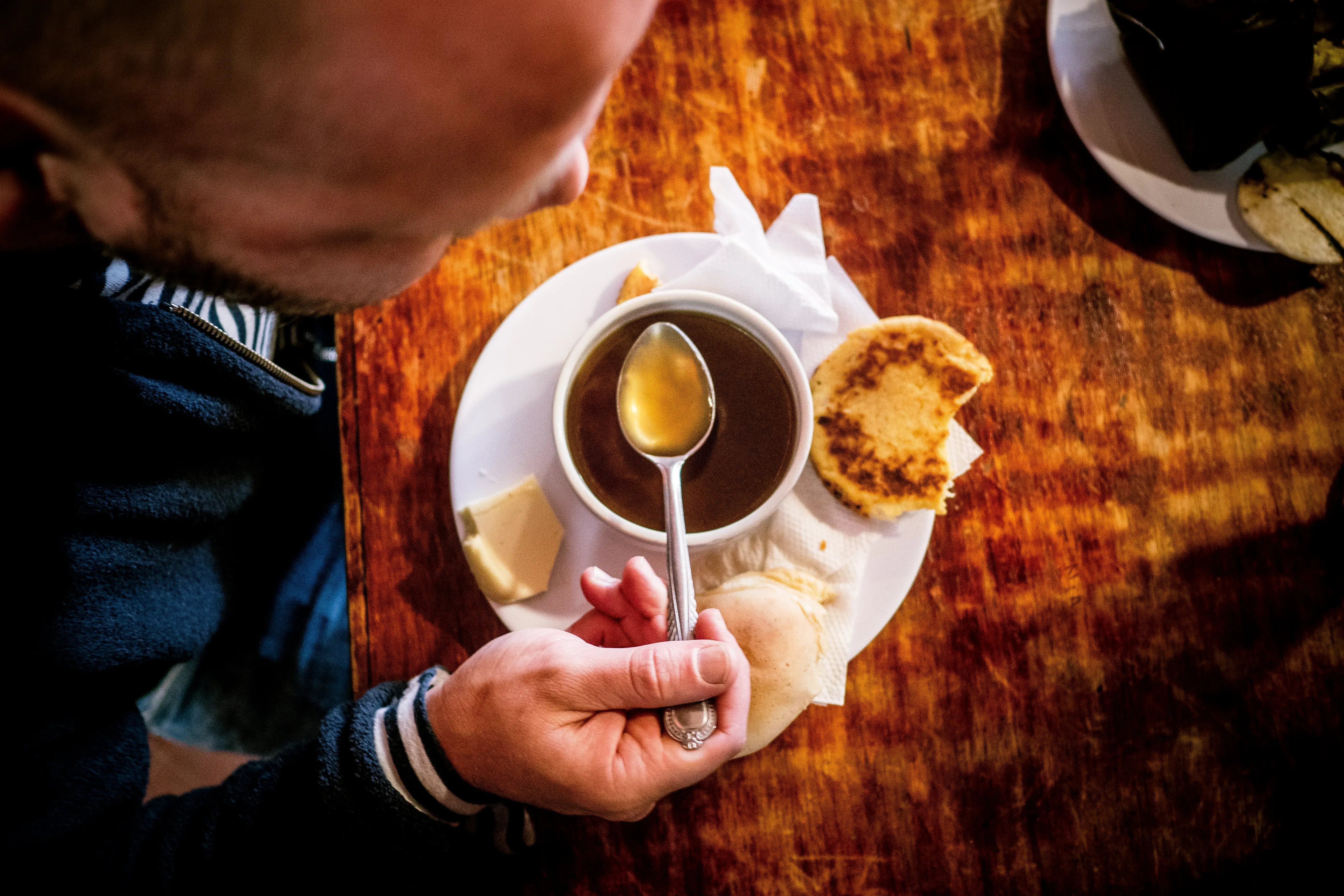 Vista superior de una persona comiendo un ajiaco o caldo con arepa y queso en una mesa rústica.