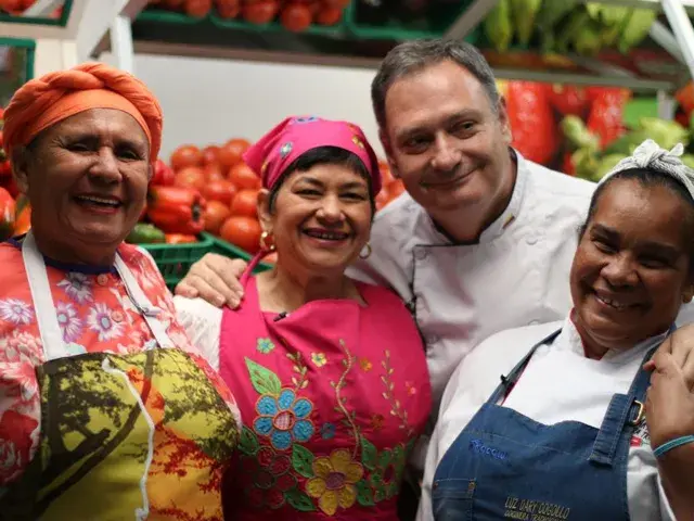 Grupo de chefs sonriendo juntos frente a un puesto de tomates frescos.