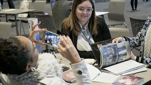 Dos mujeres en una mesa, una toma una foto con su móvil y la otra mira un folleto.