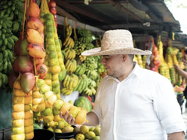 Chef en un mercado sosteniendo frutas frescas con un puesto lleno de productos.