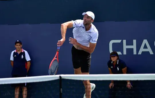 Tenista Ivo Karlovic realizando un servicio en un partido al aire libre en un estadio con fondo azul.