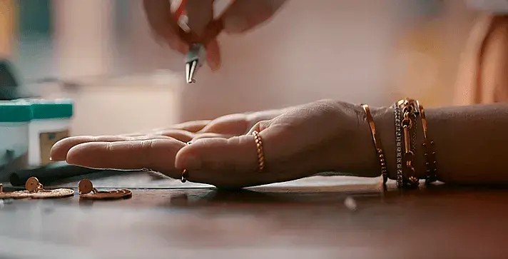 Mano con pulseras y anillos de oro, mientras una pinza trabaja en una joya.