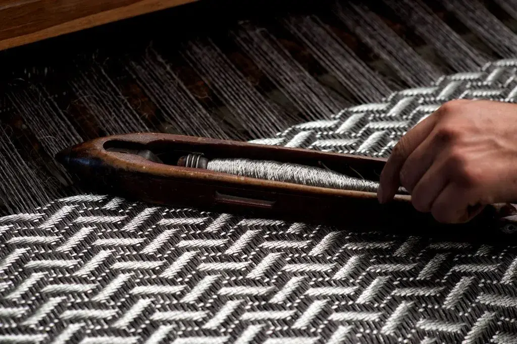 Mano teje tela gris con lanzadera y patrón zigzag en telar de madera.