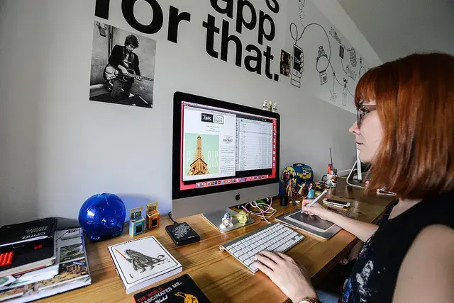 Mujer pelirroja con gafas sentada en su escritorio, trabajando en un monitor Apple.