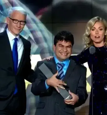 Anderson Cooper, un hombre sonriente sosteniendo un premio y Kelly Ripa en un escenario de gala.