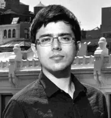 Retrato en blanco y negro de un hombre con gafas y camisa oscura frente a edificios urbanos